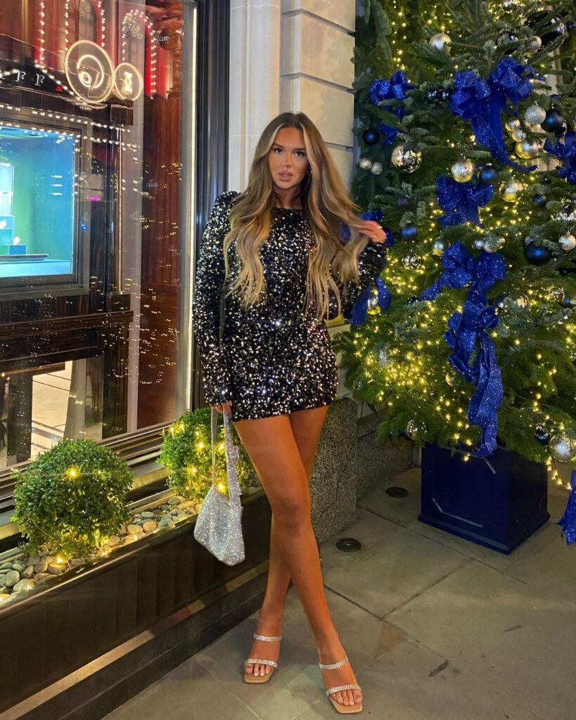 Woman in a black sequin dress standing beside a decorated Christmas tree outdoors.