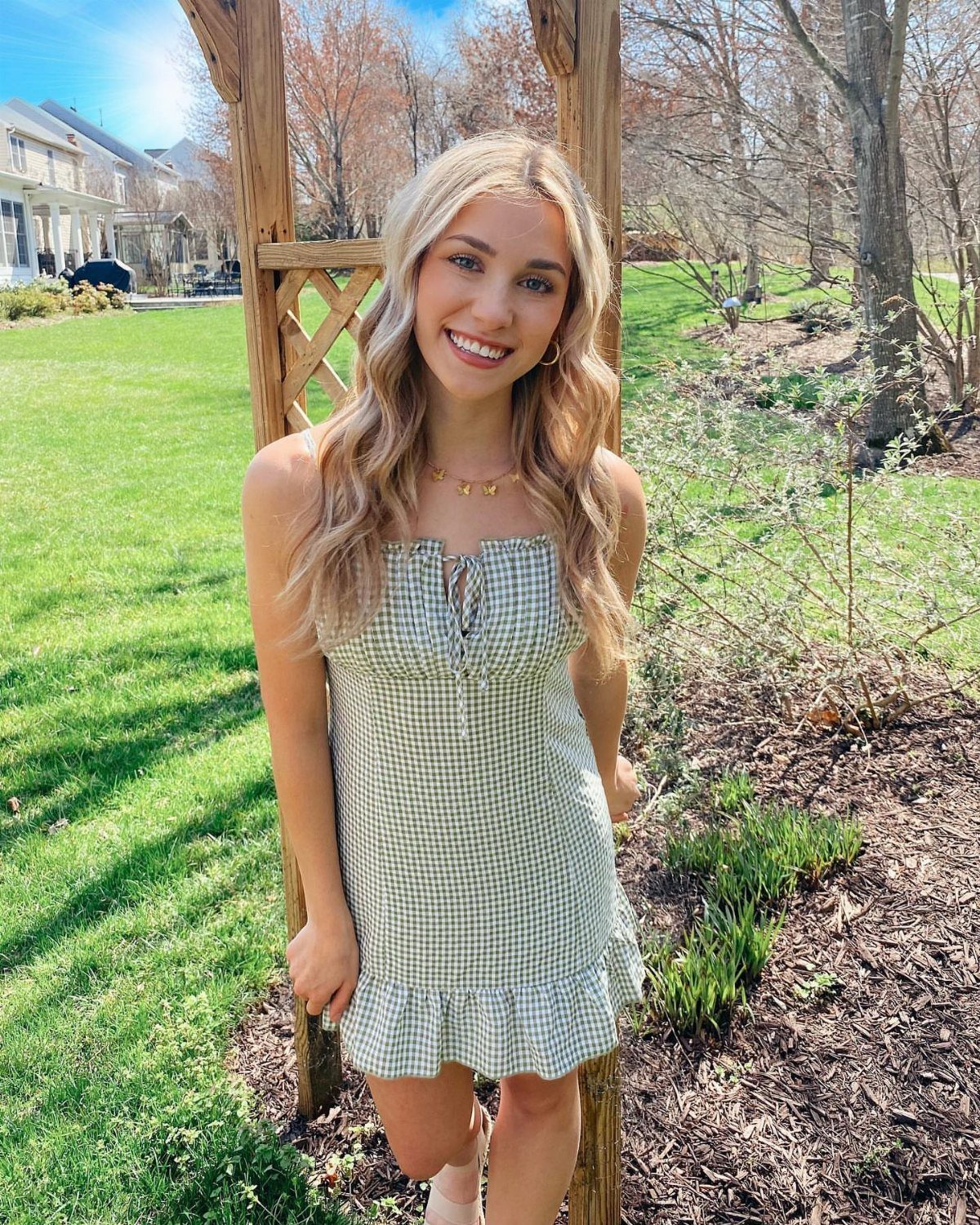 Woman in a green gingham dress smiling in front of a wooden garden trellis.