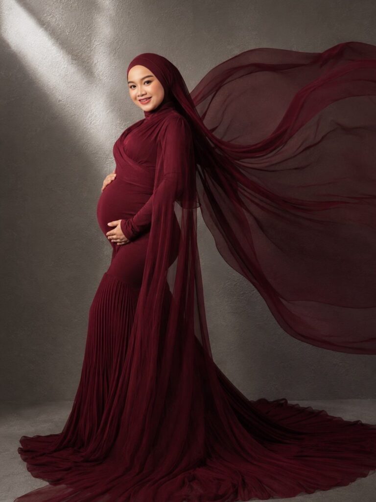 Pregnant woman in a flowing burgundy maternity gown and hijab posing in studio