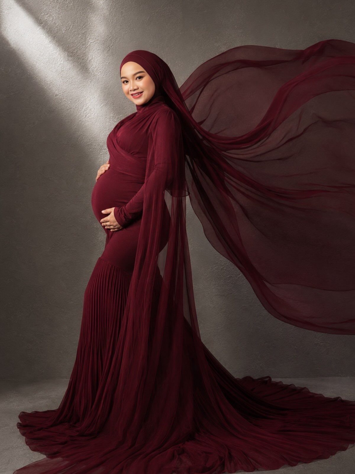 Pregnant woman in a flowing burgundy maternity gown and hijab posing in studio