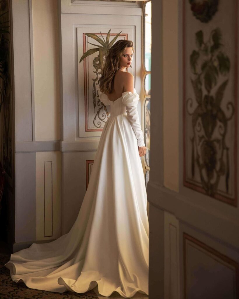 Woman in a minimalist off-the-shoulder white satin wedding dress standing in an ornate room.