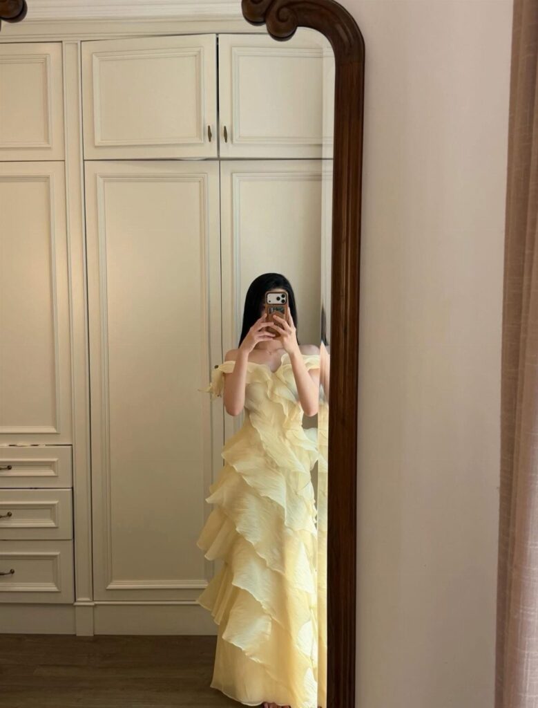 Woman in a tiered pale yellow ruffle dress taking a mirror selfie indoors.