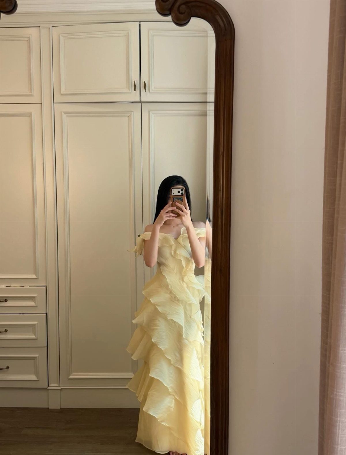 Woman in a tiered pale yellow ruffle dress taking a mirror selfie indoors.