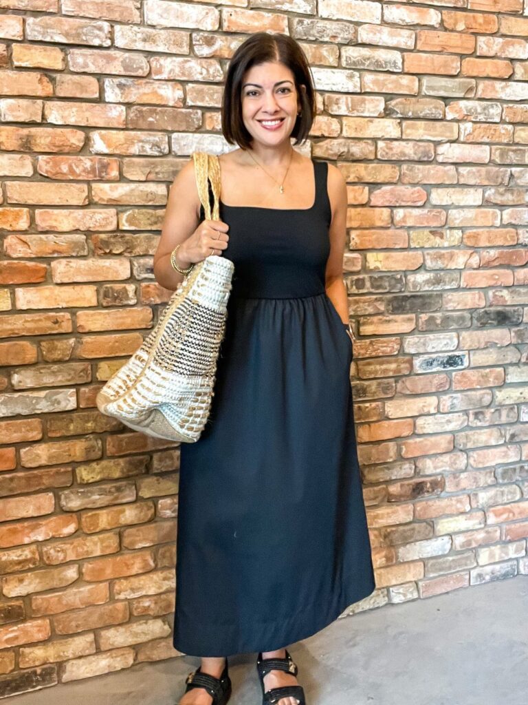Woman in a black midi tank dress holding a woven straw bag by a brick wall