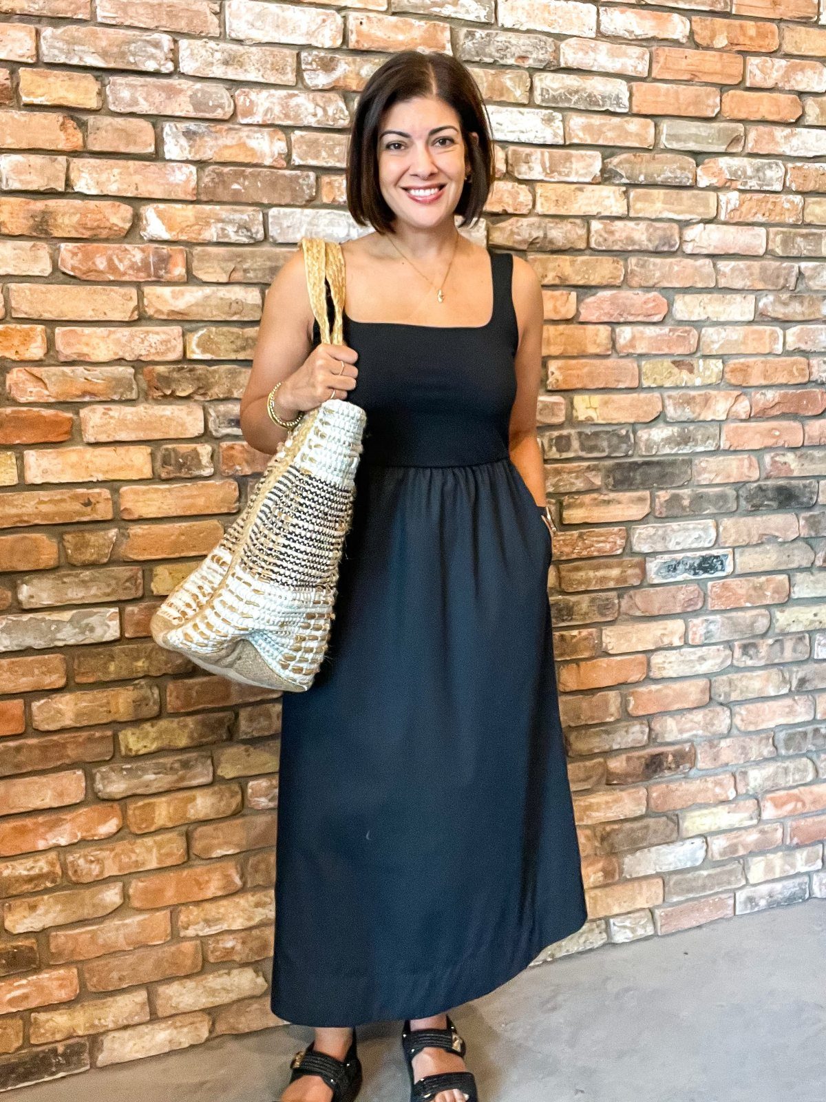 Woman in a black midi tank dress holding a woven straw bag by a brick wall