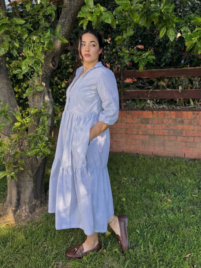 Woman wearing a light blue patterned midi shirt dress standing outdoors by a tree.