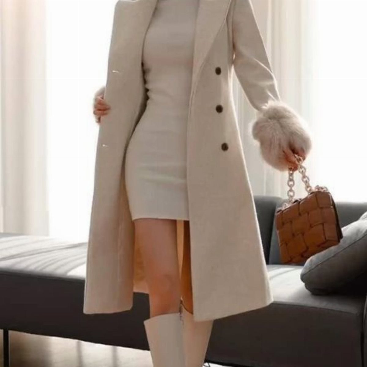 Woman in a cream dress and long matching coat with white boots and brown handbag.