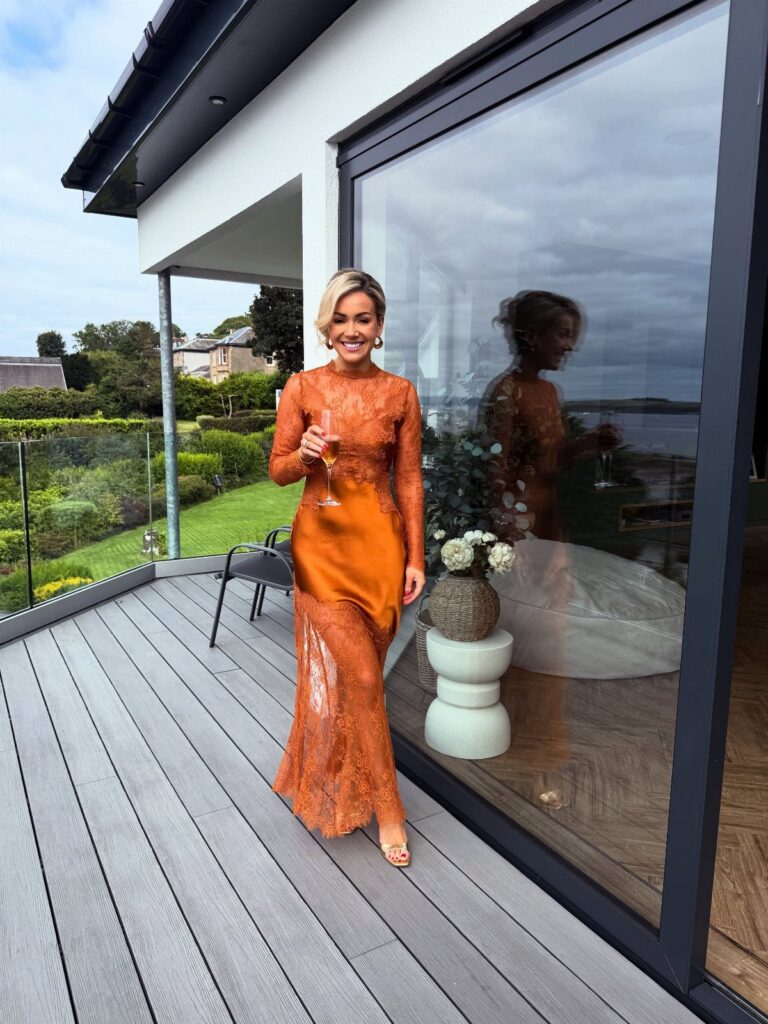 Woman in a shimmering orange party dress holding champagne on a wooden deck.